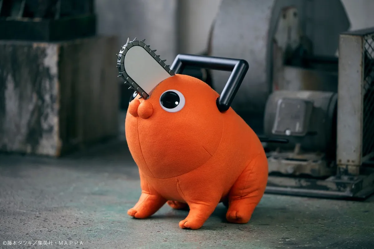 MegaHouse LIFE SIZE DOLL Pochita from the TV anime Chainsaw Man, a round orange dog-like devil with a chainsaw blade sticking out of its head, a black handle on its back and large round eyes, sitting on a concrete floor in front of industrial machinery, official product photo