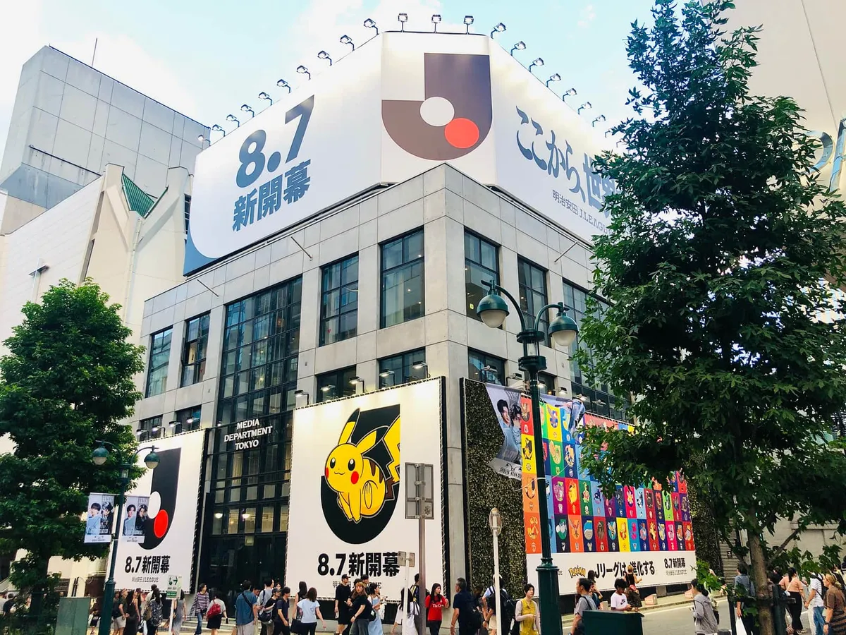 Shibuya PARCO seen from the street corner in August 2026, with a Pikachu and J.League tie-up banner on the facade and shoppers on the sidewalk